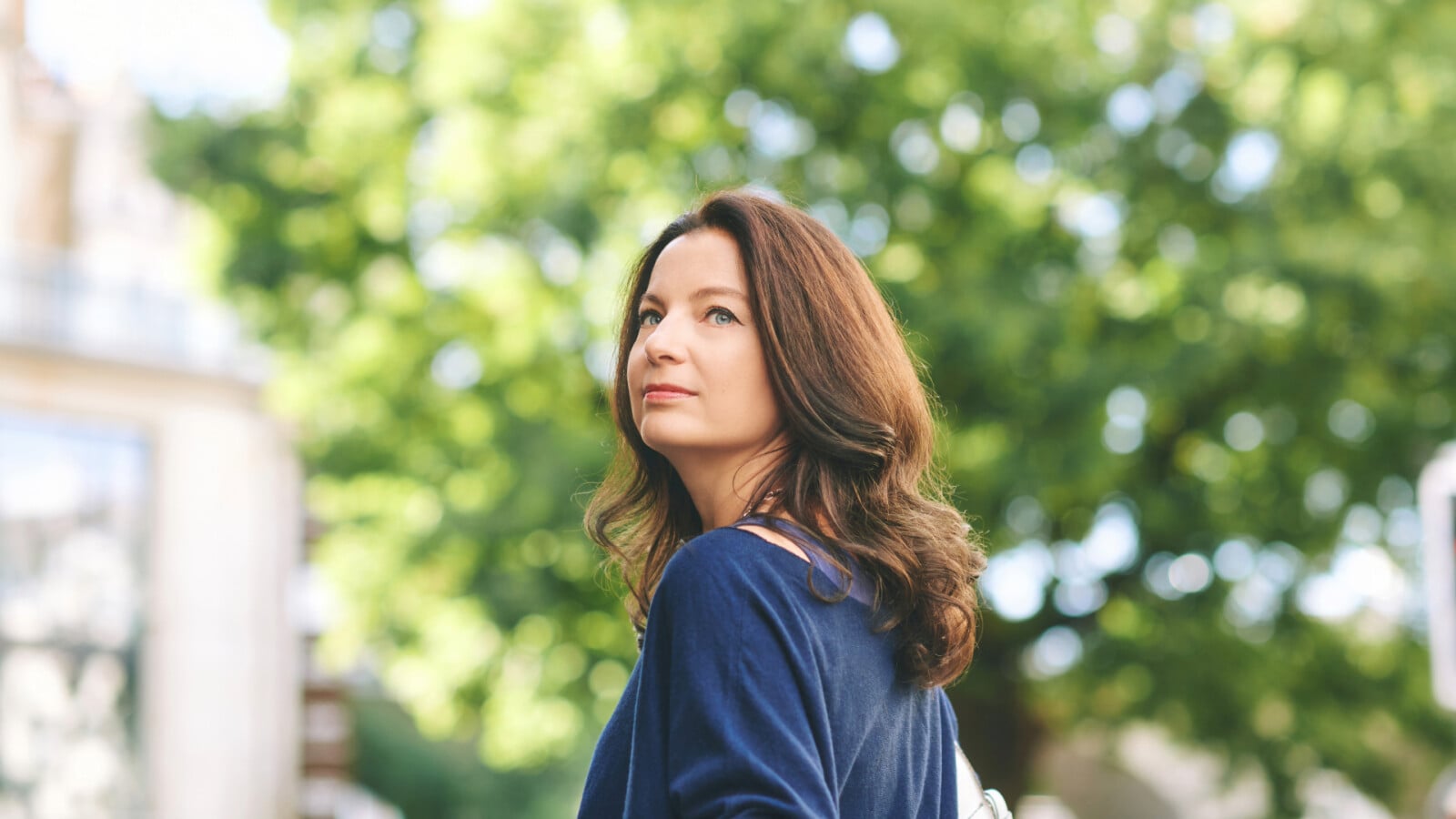 A woman with long brown hair stands outdoors in front of green trees, looking thoughtfully into the distance on a sunny day